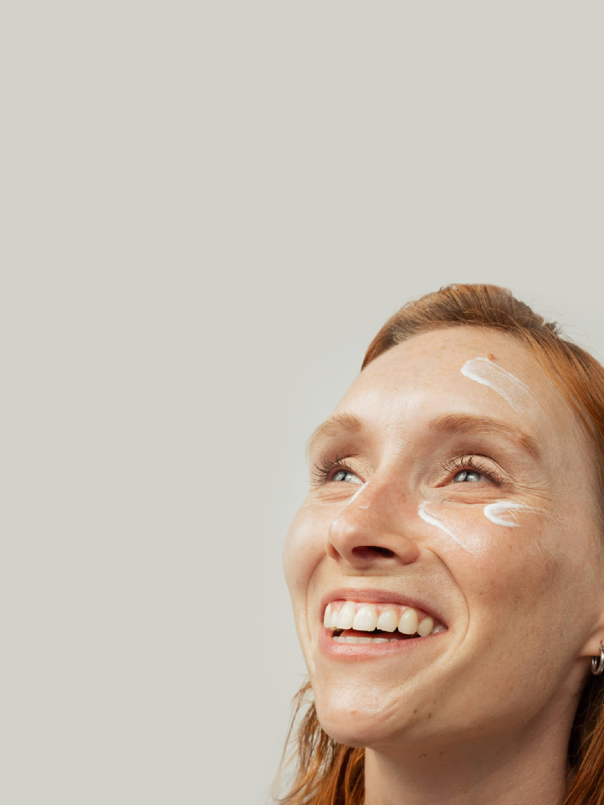 Portrait d’une femme souriante avec des touches de crème appliquées autour des yeux, sur fond neutre.